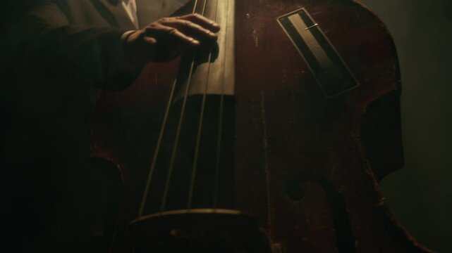 Close up shot of adult man playing upright bass on stage in spotlight with focus on hands strumming strings and spinning instrument for dramatic flare, copy space