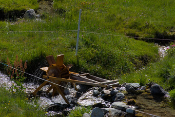 A small, rustic wooden water wheel stands beside a flowing stream in a vibrant green landscape, evoking a sense of traditional charm, peaceful nature, and sustainable simplicity.