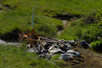 A small, rustic wooden water wheel stands beside a flowing stream in a vibrant green landscape,...