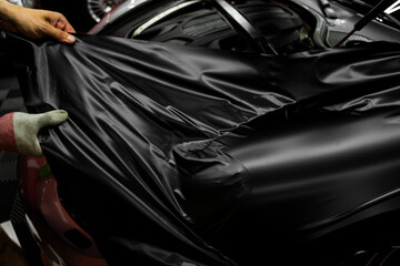 Car wrapping process using black matte vinyl stretched into place. Careful application and tensioning of black vinyl avoids surface damage or bubbling. Matte black vinyl is applied across car hood.