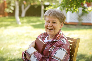 Smiling happy senior woman in plaid jacket holding book while sitting on wooden chair outdoors on a sunny day, concept of happiness, theme of aging, reading, relaxation, retirement