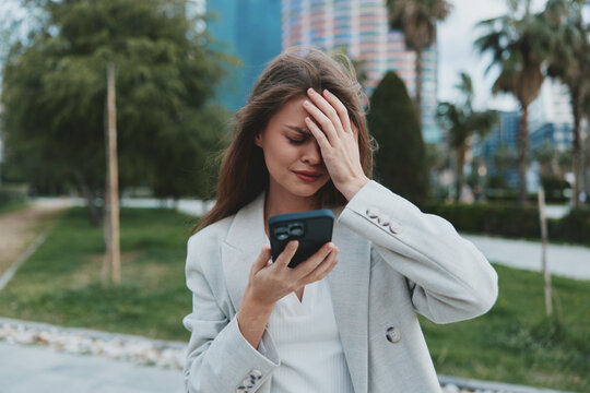 Woman stressed by financial crisis reading stock market fall news on smartphone in urban outdoor setting with green palms and buildings - Powered by Adobe