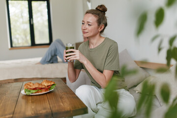 Middle-aged woman drinking green smoothie indoors, showcasing healthy lifestyle, wellbeing, active...