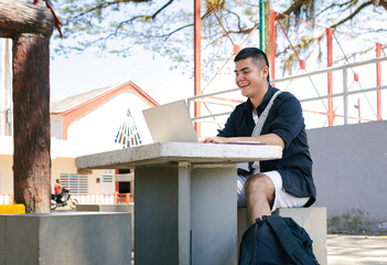 A young Latino man smiles happily while using his laptop at an outdoor concrete table, working as a...