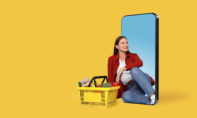 Woman with shopping basket full of products sitting in mobile phone against orange background....