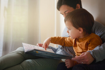 Toddler pointing at a page while reading with his mother in bed