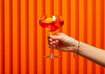 Elegance in a glass: A classic Aperol Spritz, poised in a woman's hand, offering a refreshing escape on a warm, inviting orange canvas.