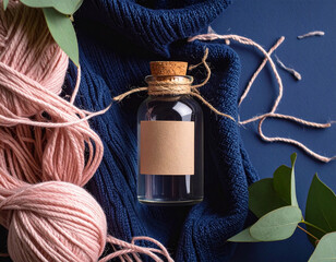 Glass bottle with blank label on blue fabric surrounded by pink yarn and scattered green leaves