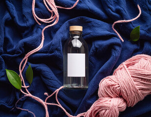 Glass bottle with blank label on blue fabric surrounded by pink yarn and scattered green leaves