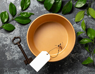 Clay bowl with blank tag and antique key surrounded by green leaves on dark textured surface