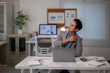 A man in a suit is sitting at a desk with a laptop and a stack of papers