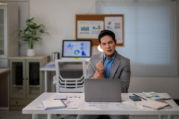 A man in a suit is sitting at a desk with a laptop and a stack of papers