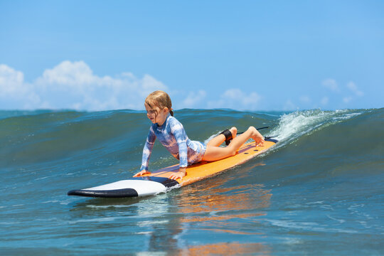 Happy baby girl - young surfer ride on surfboard with fun on sea waves. Active family lifestyle, kids outdoor water sport lessons and swimming activity in surf camp. Beach summer vacation with child.