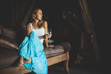 Obraz premium Young woman sit on sofa inside luxury wooden villa. Girl relaxing on family summer vacation. Tropical lifestyle.