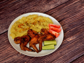 Baked chicken wings with spices, mashed potatoes, tomatoes and cucumber on a wooden table.
