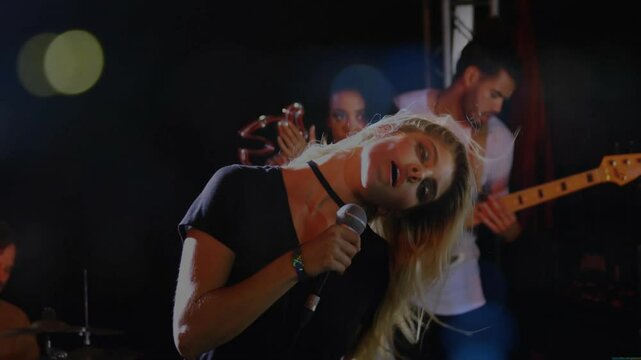 Singer gripping microphone and singing on stage, bandmates playing guitar and drums in bokeh lights