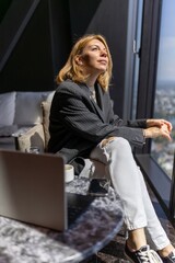 A stylish woman in a blazer sits by a large window in a modern office, enjoying the city view. This scene emphasizes business, productivity, and the chic lifestyle of a modern businesswoman