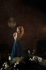 Young Caucasian woman in blue dress posing with raised arms, looking away. Front view. Vintage room with brick wall, crystal chandelier, flowers. Concepts: elegance, beauty, nostalgia, romance.