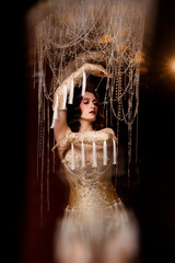 Elegant young circus actress in golden corset and gloves, standing under chandelier, front view. Retro theatrical look for circus, burlesque, performance, vintage stage shows.