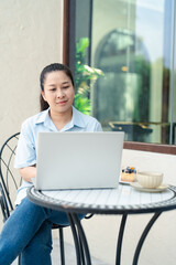 Asian woman working on laptop outdoors at a cafe table, enjoying coffee and dessert in peaceful environment, concept of remote work, freelance lifestyle, digital nomad and modern productivity setting.