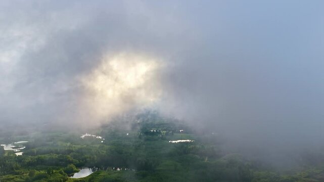 A cinematic drone shot pulls back just beneath a layer of dramatic, moody clouds, revealing majestic sunbeams, crepuscular rays breaking through to illuminate vast green forest and lakes of Latvia.