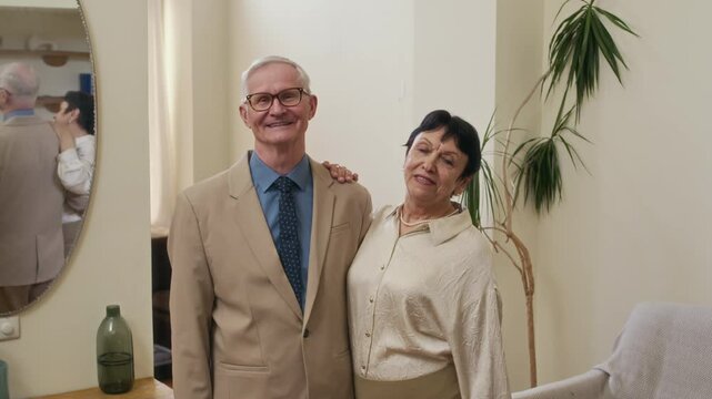 Tilt up of happy affectionate elderly spouses wearing elegant attire looking at camera with smiles while standing on wooden floor in spacious living room