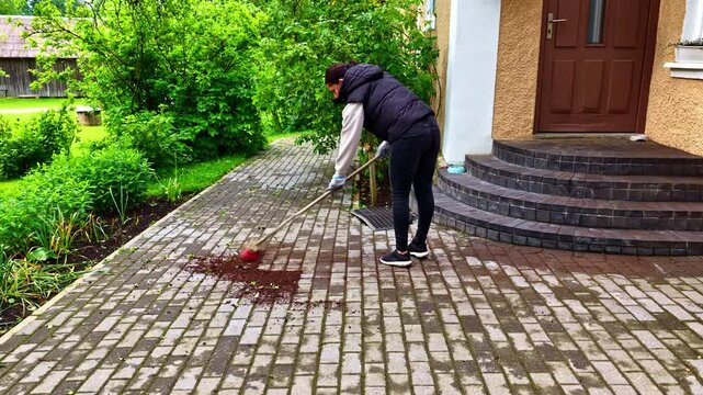 A woman does yard work, using a push broom to sweep up a pile of wet soil from the interlocking paving stones of her house's backyard patio after gardening