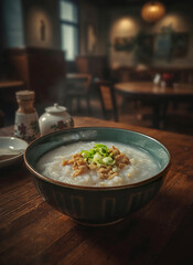 Delicious rice porridge with pork and green onions served in a ceramic bowl on a wooden table, with a blurred restaurant background.