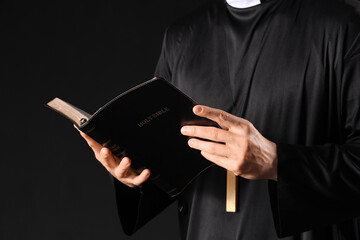Young priest reading Holy Bible on black background, closeup