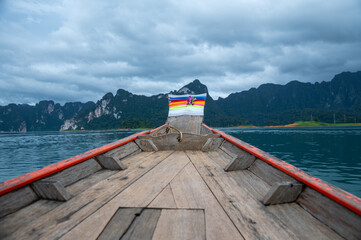 Wooden long-tail boat cruising on a calm lake with lush green mountains and cloudy sky in the...