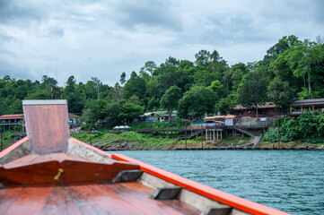 Wooden long-tail boat cruising on a calm lake with lush green mountains and cloudy sky in the...