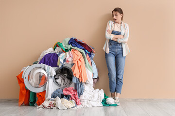 Troubled young woman near washing machine with pile of dirty laundry on beige background