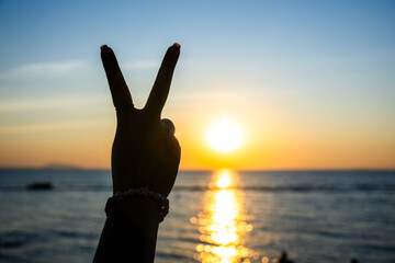 silhouetted hand making a peace sign against a glowing sunset over the ocean, with golden light