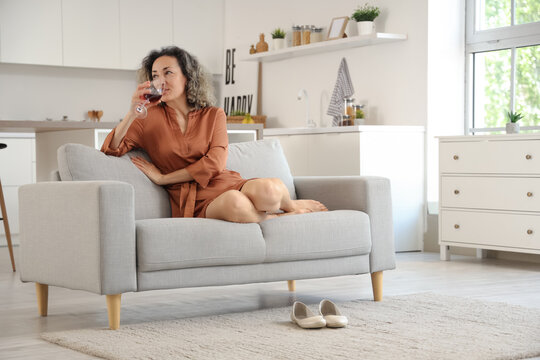 Beautiful middle-aged woman with glass of red wine sitting on sofa at home