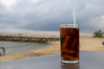 glass of iced cola sits on a table by the sea, with a cloudy sky backdrop. The drink's bubbles and condensation are visible