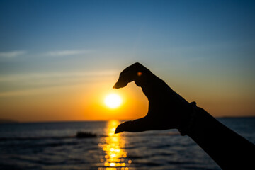 silhouetted hand forming a half-heart gesture in front of a glowing sunset over the ocean