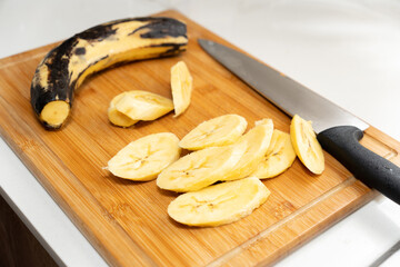 Photograph of a wooden chopping board with bananas cut with a knife on a white table.
