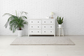 Chest of drawers, carpet and houseplants near white brick wall in living room