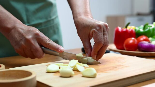 Closeup of hands slicing fresh garlic on a wooden cutting board in kitchen. Perfect for cooking, food prep, healthy eating, herbs