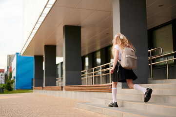 Obraz premium schoolgirl with backpack climbs steps of modern building. education concept with young student heading to school on sunny day. urban architecture enhances academic theme
