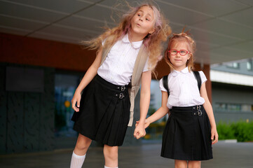 Obraz premium schoolgirls in uniform holding hands, standing outside building. young friends with backpacks, ready for school day. cute childhood moment, education concept, cheerful expression.