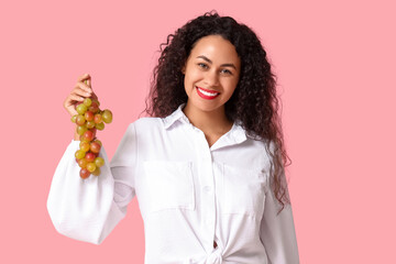 Happy young African-American woman with sweet ripe grapes on pink background