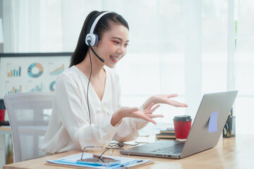 Asian woman wearing headset works at her desk in home office, engaging in online customer service and technical support, professional communication and leadership during virtual meetings and webinars