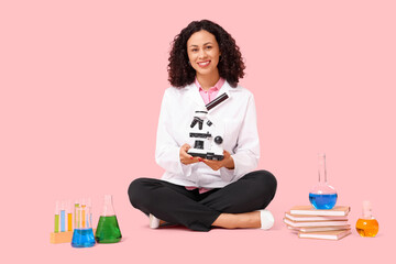 Female African-American scientist with microscope and flasks sitting on pink background