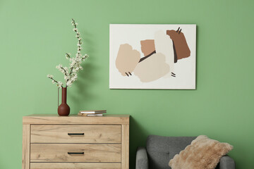 Vase with blooming tree branches and books on chest of drawers near green wall