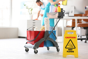 Trolley with cleaning supplies and caution sign against janitors in office