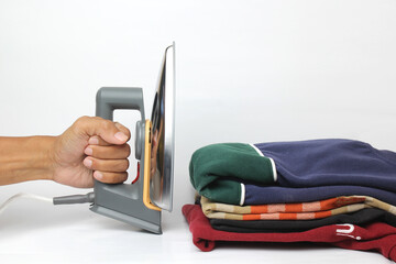 A hand holds an iron, poised to press a stack of folded, colorful clothes against a white background