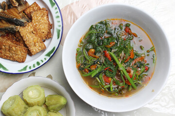A top-down view of a bowl of cooked pumpkin leaves (masakan daun labu kuning), accompanied by fried tempeh and bitter melon