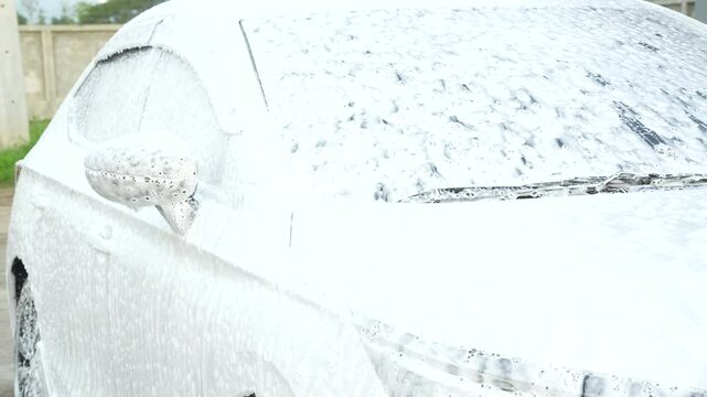A middle-aged Asian man carefully washes his car using water and foam. Each step from high-pressure rinsing to scrubbing and drying ensures the vehicle is spotless, well-maintained, and presentable