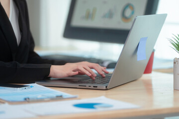 Attractive Asian woman in a formal black suit works attentively at her desk, writing notes. She embodies professionalism and focus while managing tasks in a well-organized office environment.